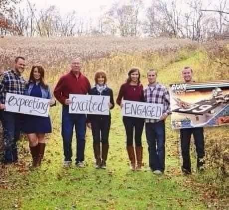 Six people stand in a grassy field holding signs that read 'expecting', 'excited', 'ENGAGED', and one person holds a 'GIANT'…