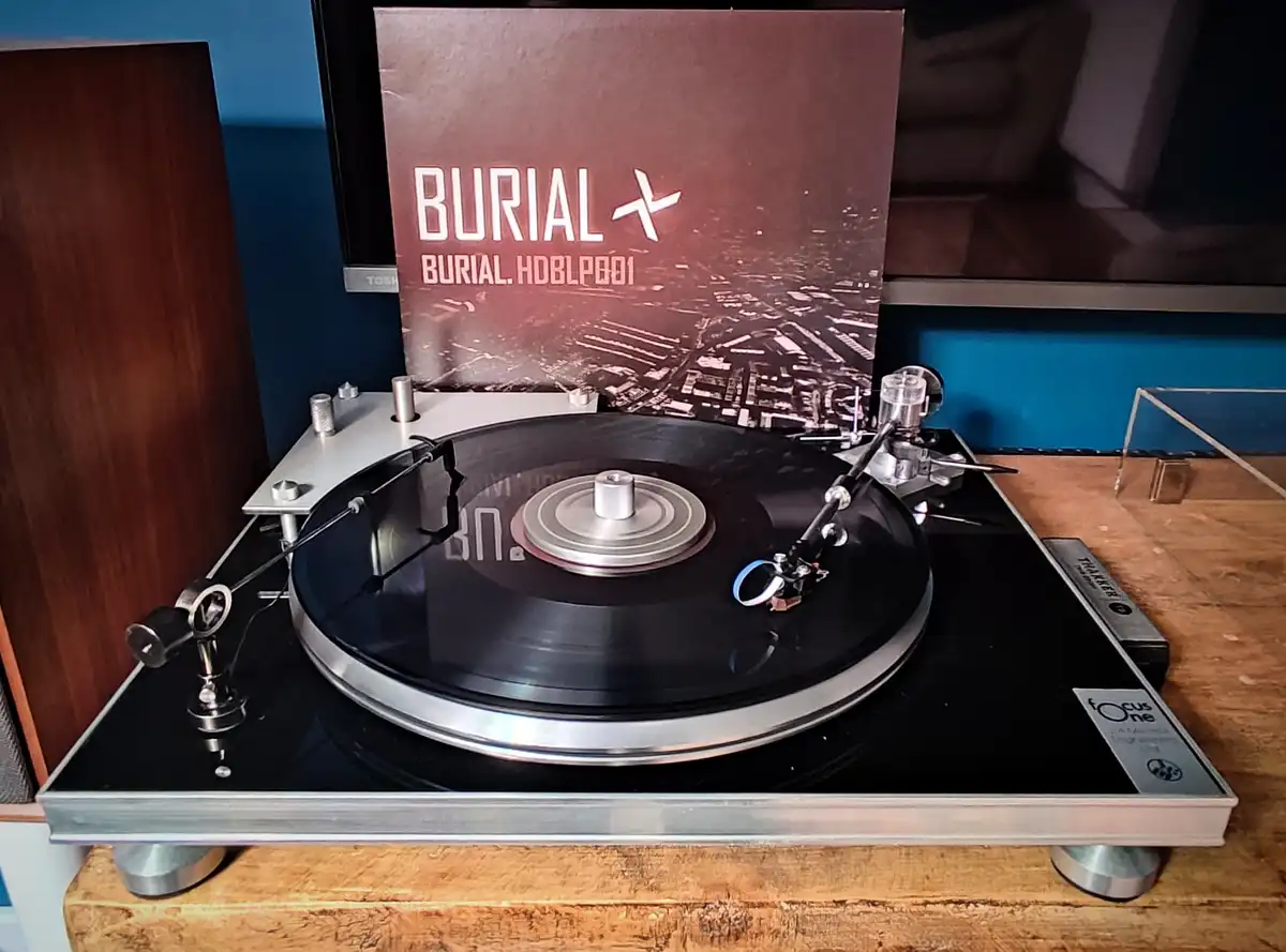 A close-up shot of a silver and black turntable with a vinyl record spinning, positioned on a wooden surface. Behind the…