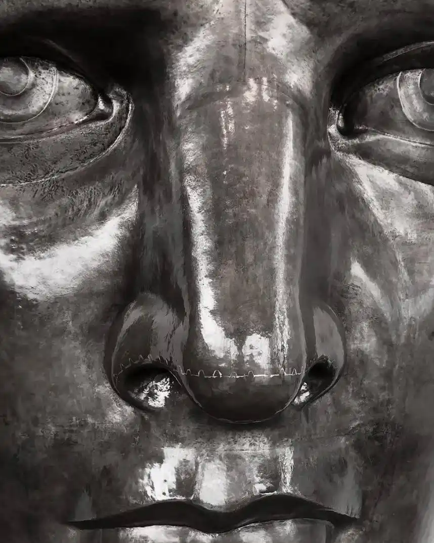 A close-up of a metallic statue's face, focusing on the nose which has visible white stitching across its bridge, suggesting…