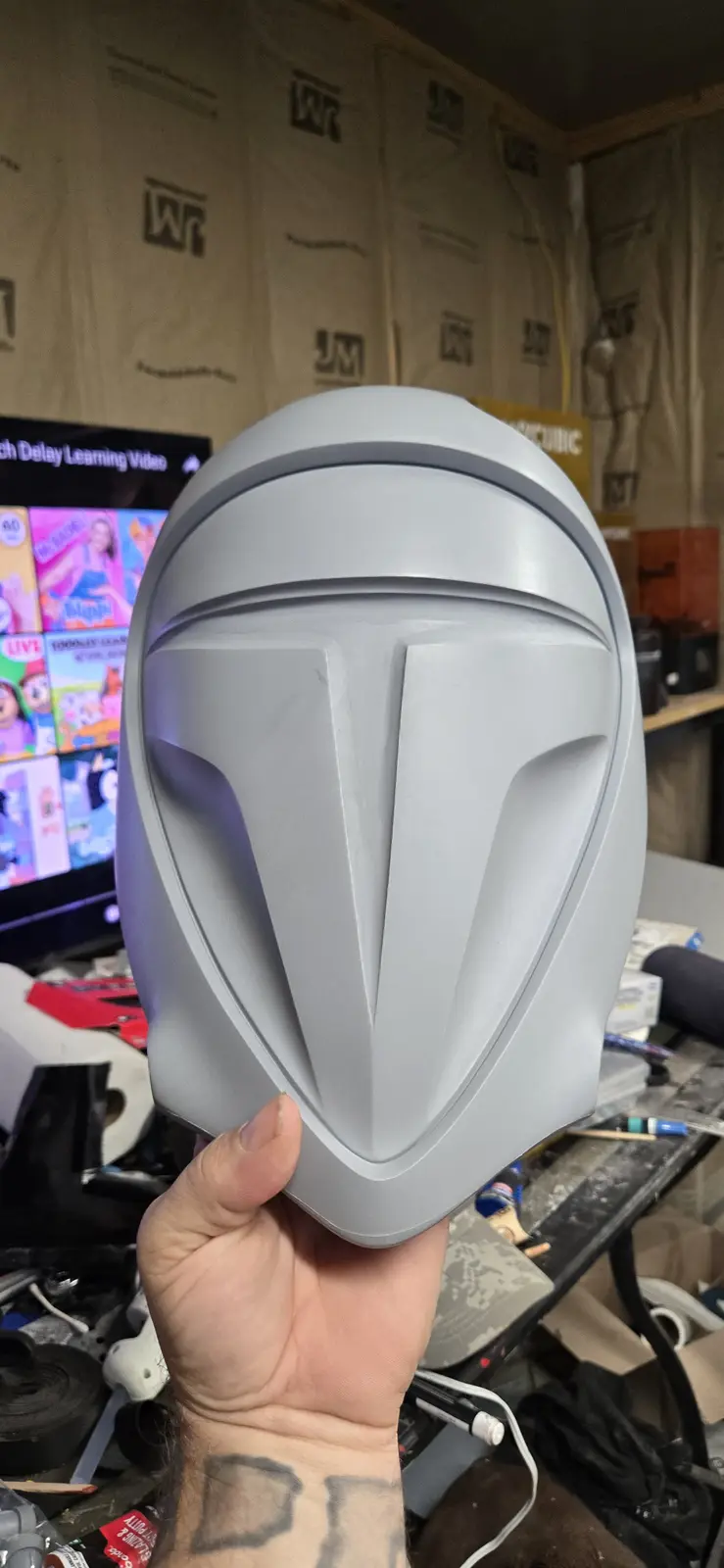 A person holds up a gray, unpainted Mandalorian-style helmet in a cluttered workshop. In the background, a TV screen…