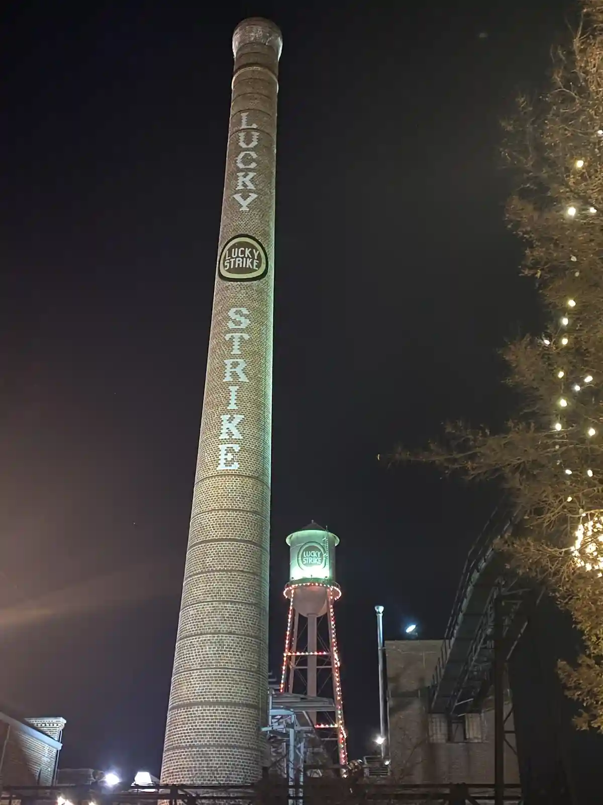 A tall brick chimney at night is illuminated with the words 'LUCKY' and 'STRIKE' vertically, with a 'LUCKY STRIKE' logo in…