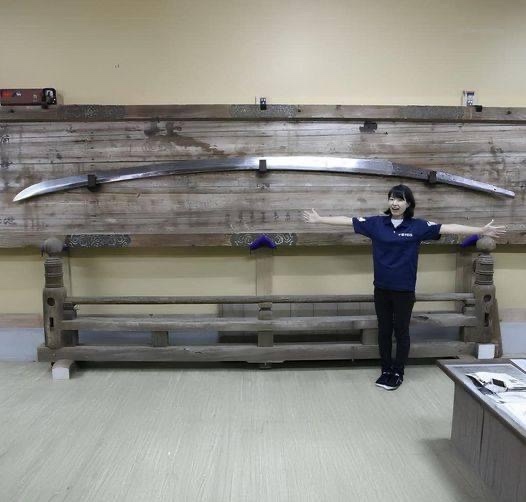 a person in a blue shirt stands with arms outstretched in front of a massive, curved sword mounted on a wooden display…