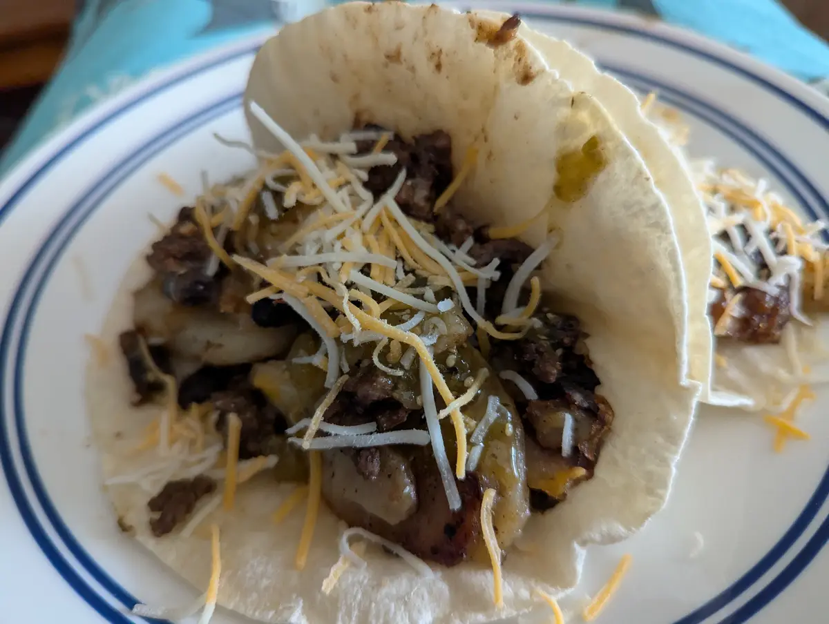 A close-up photo of a soft taco on a white plate with blue rim, filled with meat, beans, green sauce, and shredded cheese…