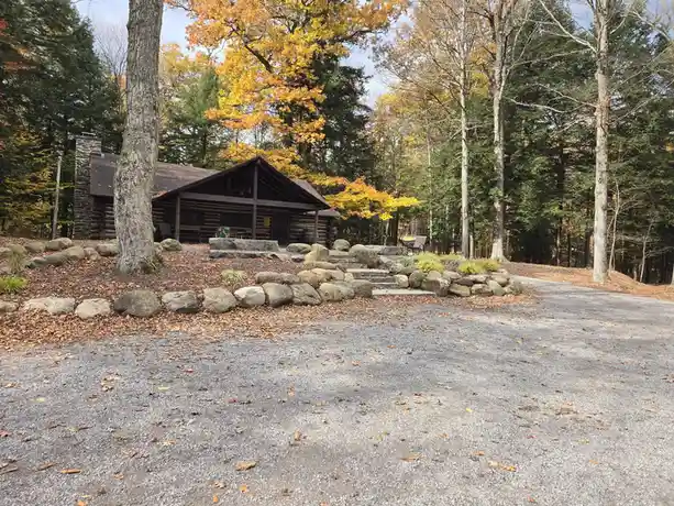 A tranquil autumn scene shows a log cabin nestled among trees with yellow and green foliage, surrounded by large rocks and a…
