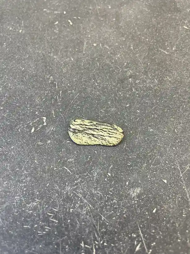 A small, elongated, pale yellowish rock with dark, wavy stripes lies centered on a heavily scratched, dark gray metallic…