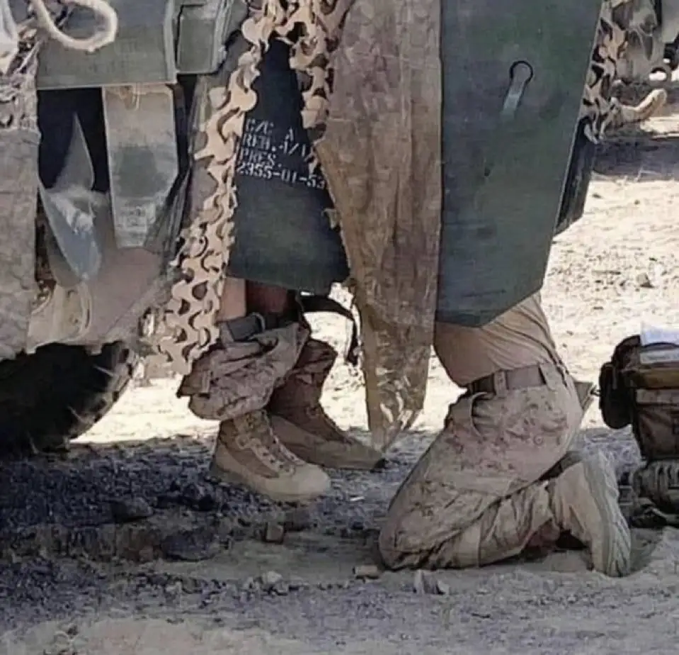 A low-angle photo shows two soldiers in desert camouflage gear and boots near a military vehicle; one is kneeling on the…
