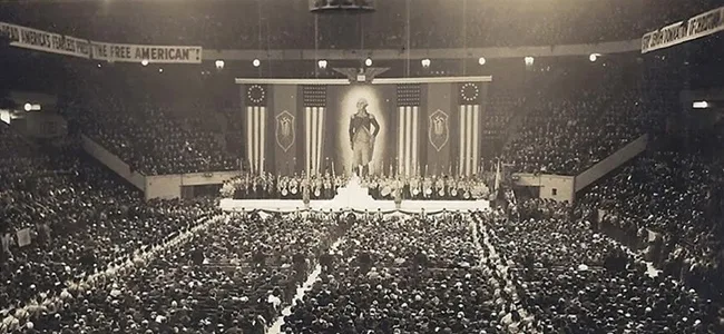 A black-and-white photo of a massive indoor rally with a large crowd seated in tiered stands facing a central stage. On the…