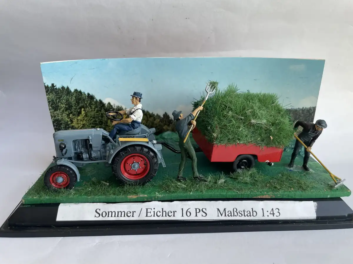 A 1:43 scale model diorama depicts a grey tractor with red wheels pulling a red trailer overflowing with green hay, while…