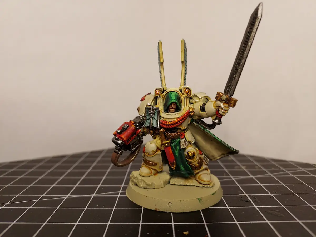 A painted miniature of a Space Marine from Warhammer 40,000 stands on a grid mat, holding a sword and a weapon, with…
