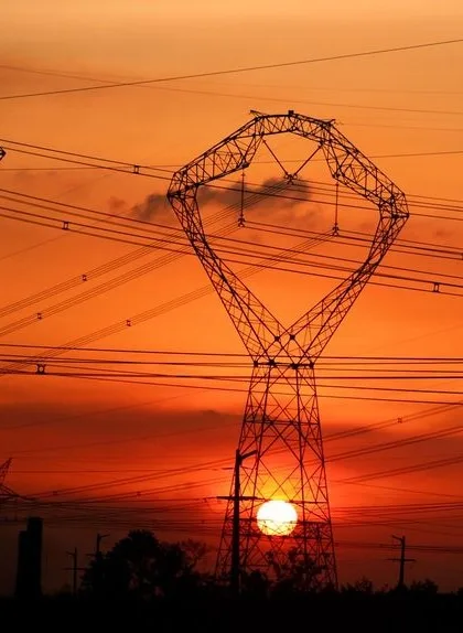 A silhouette of a large electricity pylon against a vibrant orange sunset, with the sun positioned behind it. The pylon's…