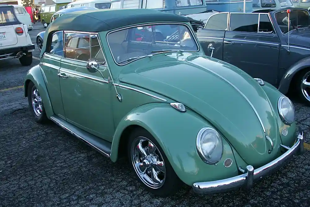 A green vintage Volkswagen Beetle with a black soft top is parked in a lot surrounded by other classic cars. The car has…