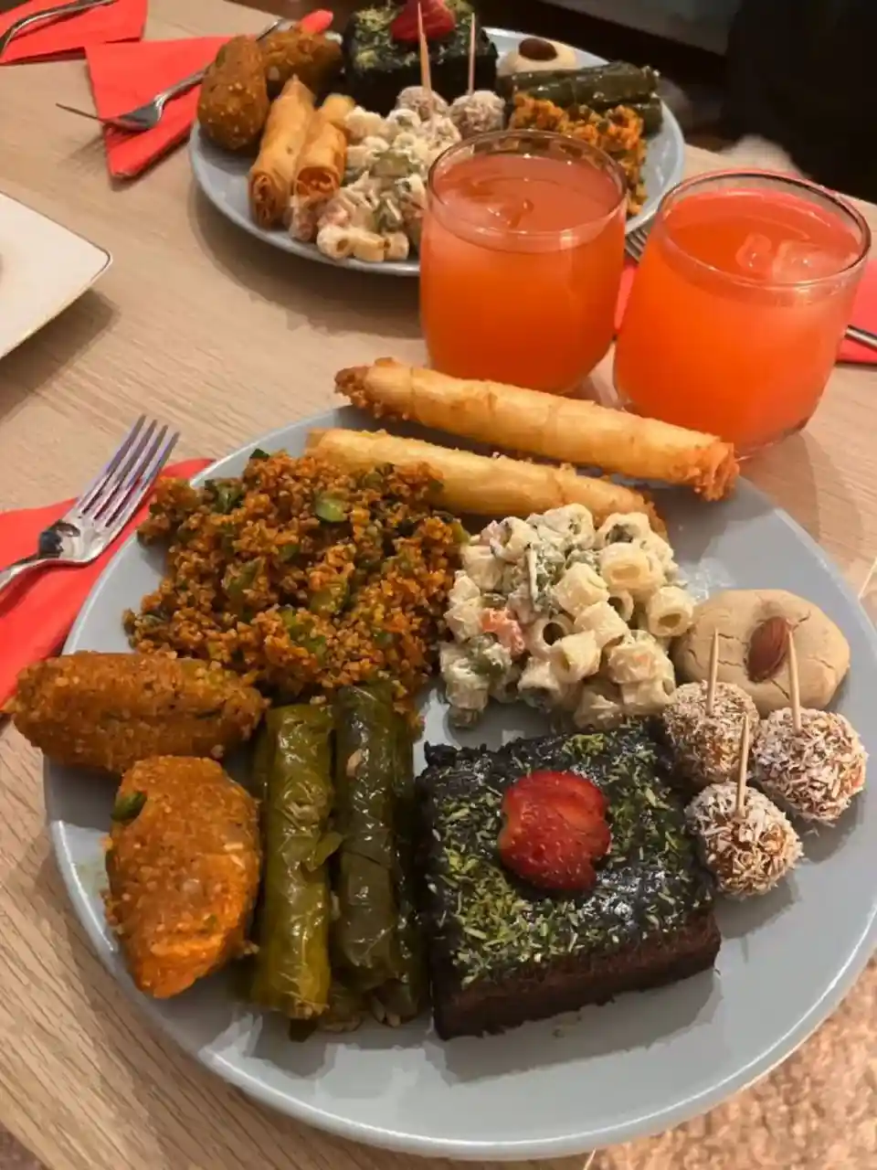A large plate of diverse, colorful food including fried rolls, pasta salad, dolmas, a dark chocolate cake with a strawberry…