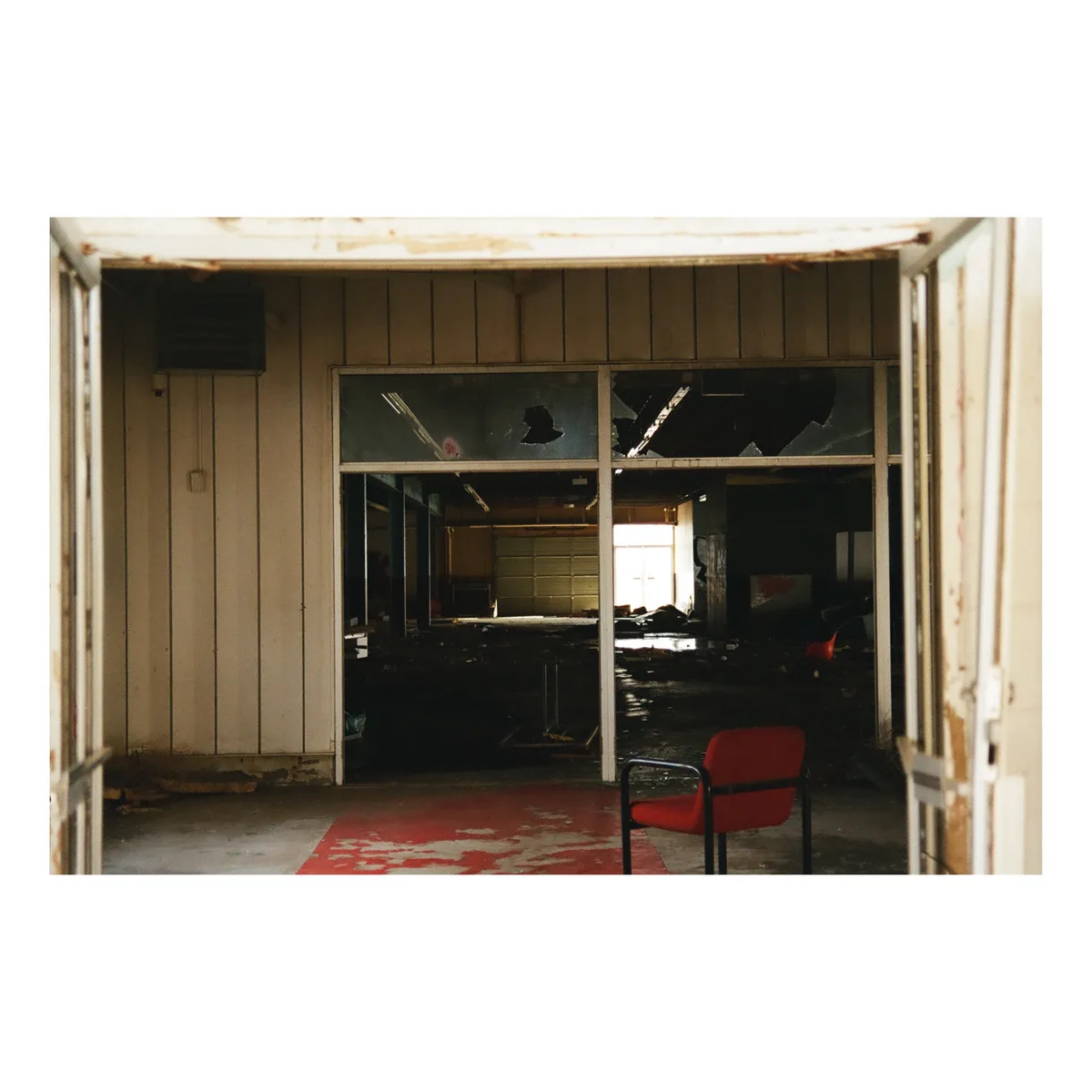 A photograph of a dilapidated, abandoned interior space viewed through an open doorway. The scene features broken glass…