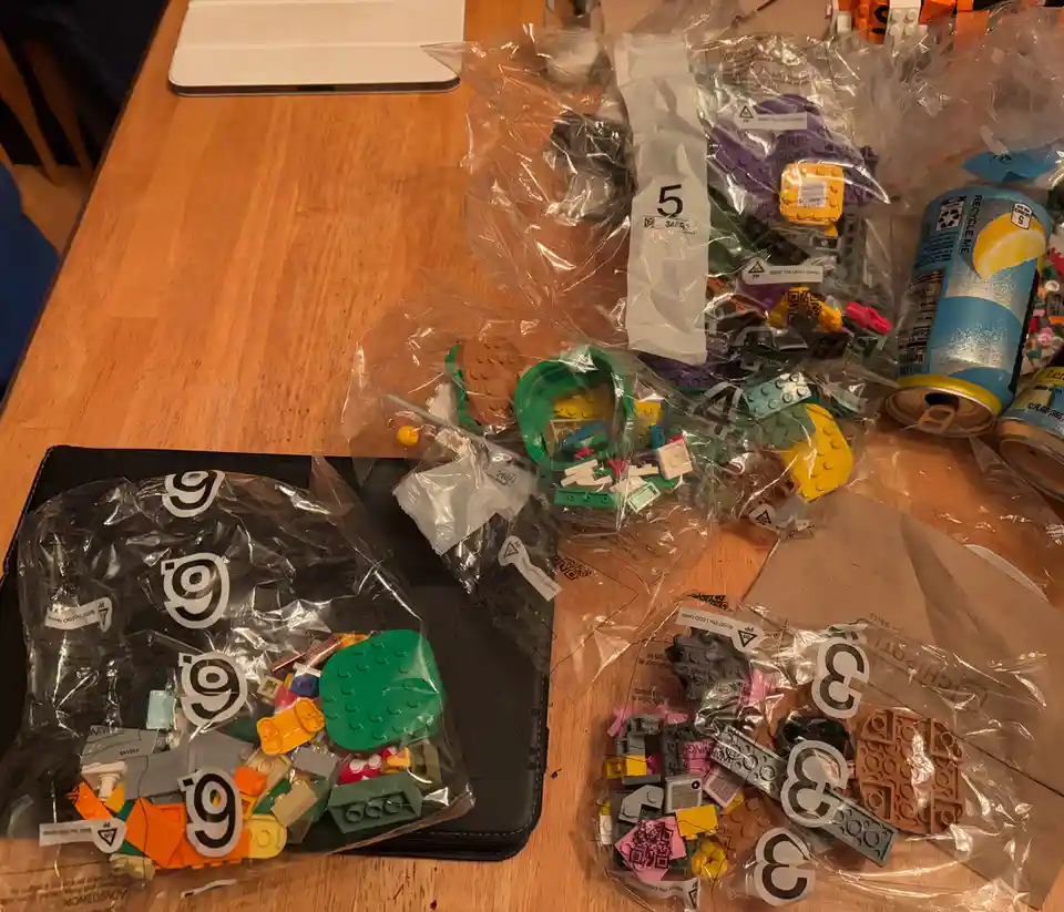 A wooden table is covered with several clear plastic bags filled with assorted Lego pieces, each bag labeled with a large…