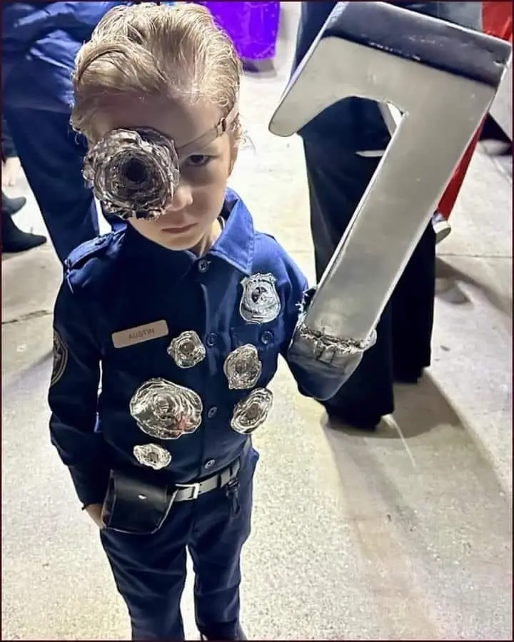 A young child dressed in a police uniform with multiple badges, holding a large metal number '7', has a large, twisted metal…
