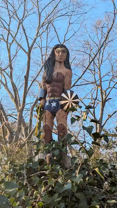 A weathered, hand-carved wooden statue of Wonder Woman stands atop a tree stump surrounded by ivy and bare trees against a…