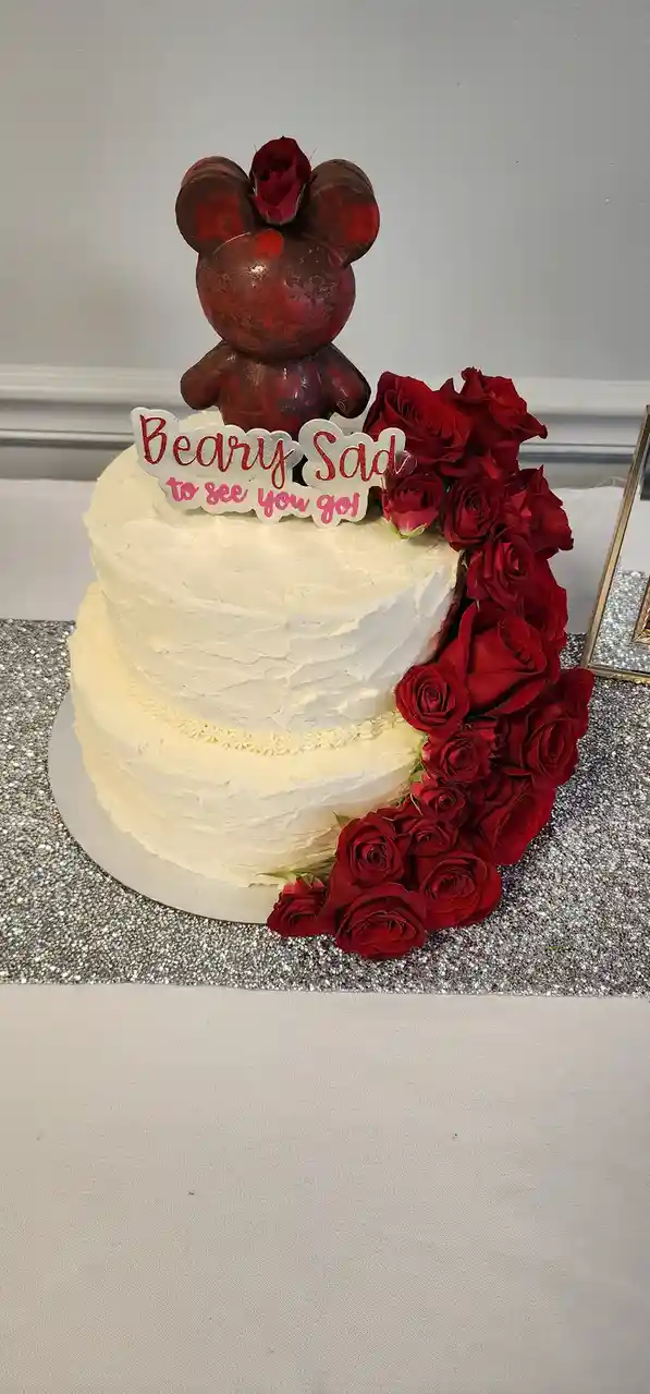 A two-tiered white frosted cake decorated with a dark red Mickey Mouse-shaped bear topper and a cascade of red roses. A…