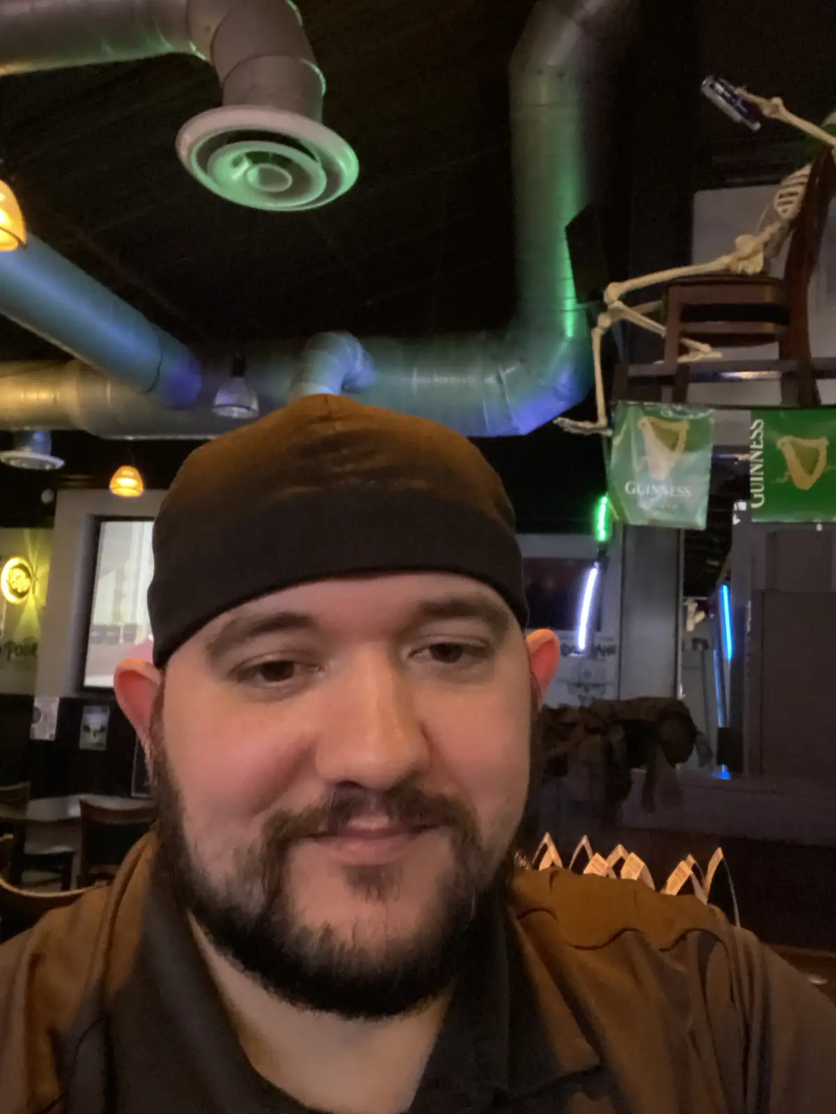 A man with a beard and a black beanie smiles at the camera in a dimly lit bar with exposed ductwork and green lighting…