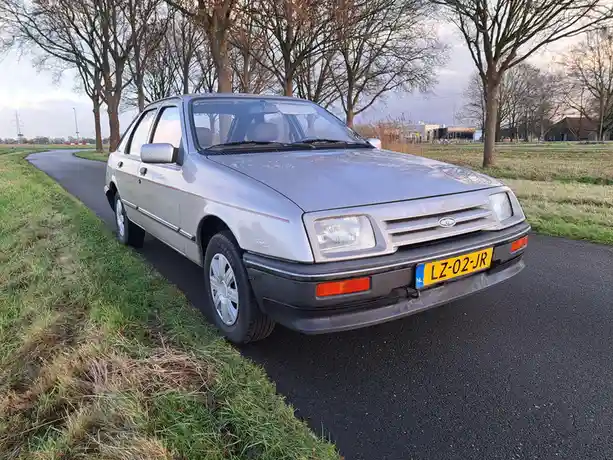 A silver 1980s Ford Escort hatchback is parked on a rural asphalt road beside grassy fields, with bare trees in the…