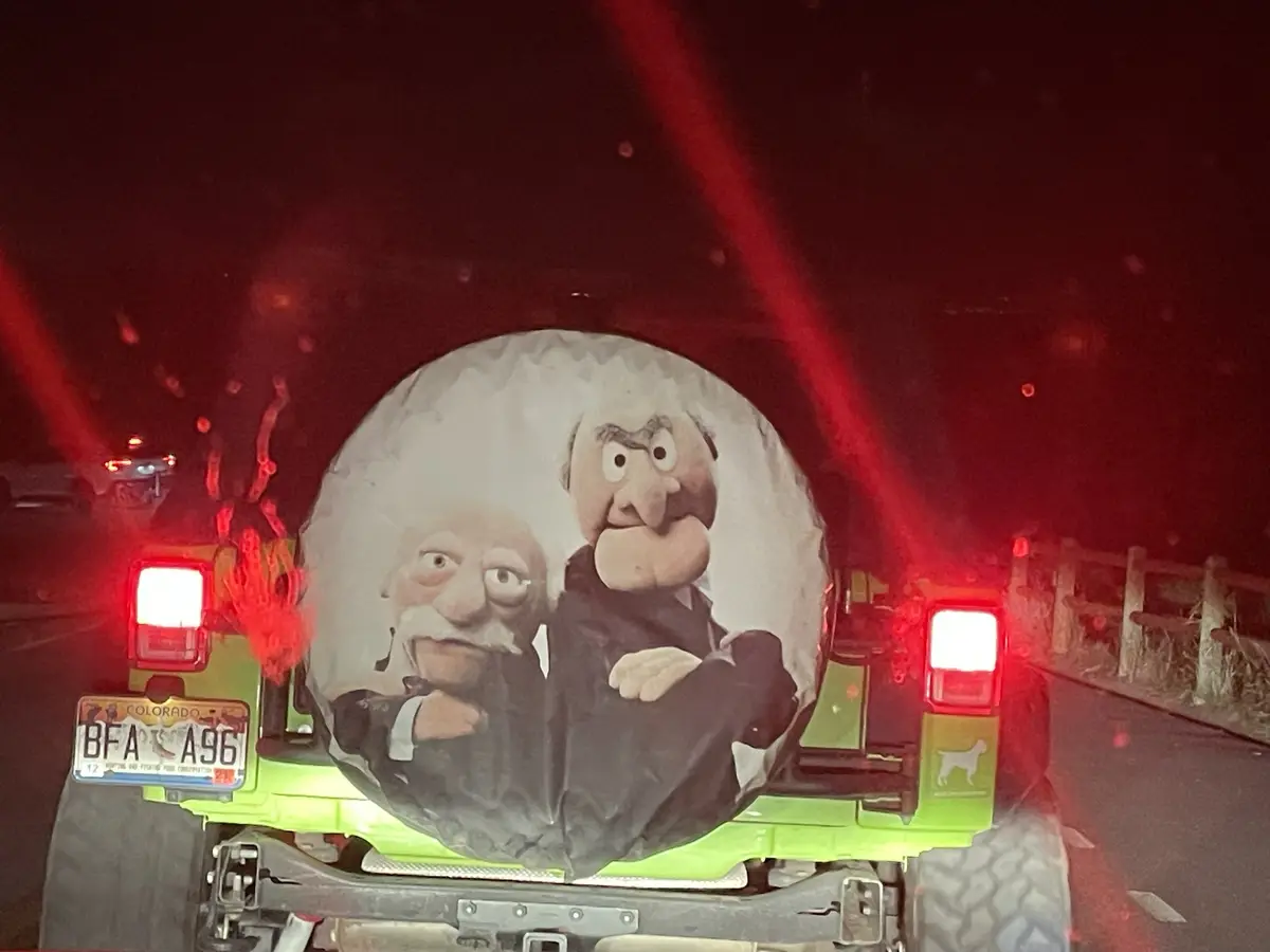 The rear of a lime-green Jeep Wrangler is shown at night, its red taillights glowing. A spare tire cover features a printed…