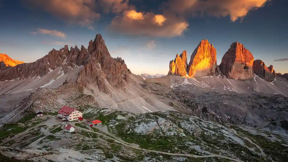 In the distance, the Three Peaks of Lavaredo, an iconic Dolomites mountain group, is bathed in a warm sunset glow. These…