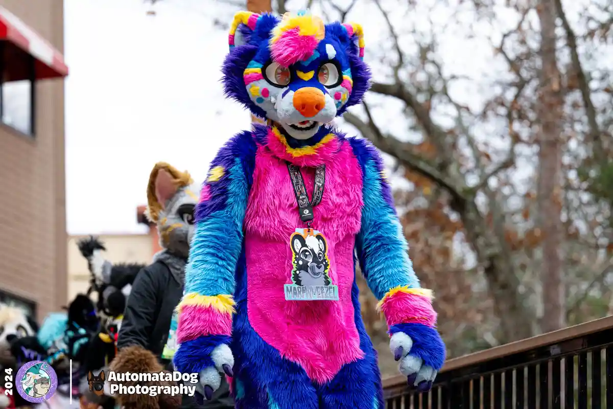 A person in a vibrant, multi-colored fursuit resembling a stylized cat or fox with blue, pink, and yellow fur, large eyes…