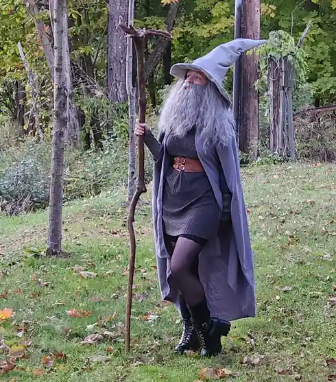 A person in a wizard costume, featuring a tall pointed hat, long grey beard, and grey robe, stands outdoors in a grassy area…