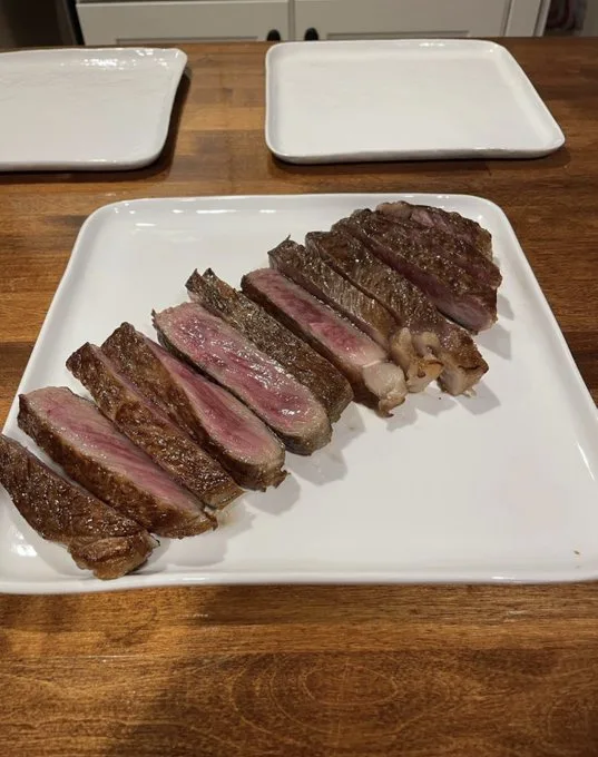 A white plate holds sliced medium-rare steak with visible pink center and seared edges, resting on a wooden table with two…