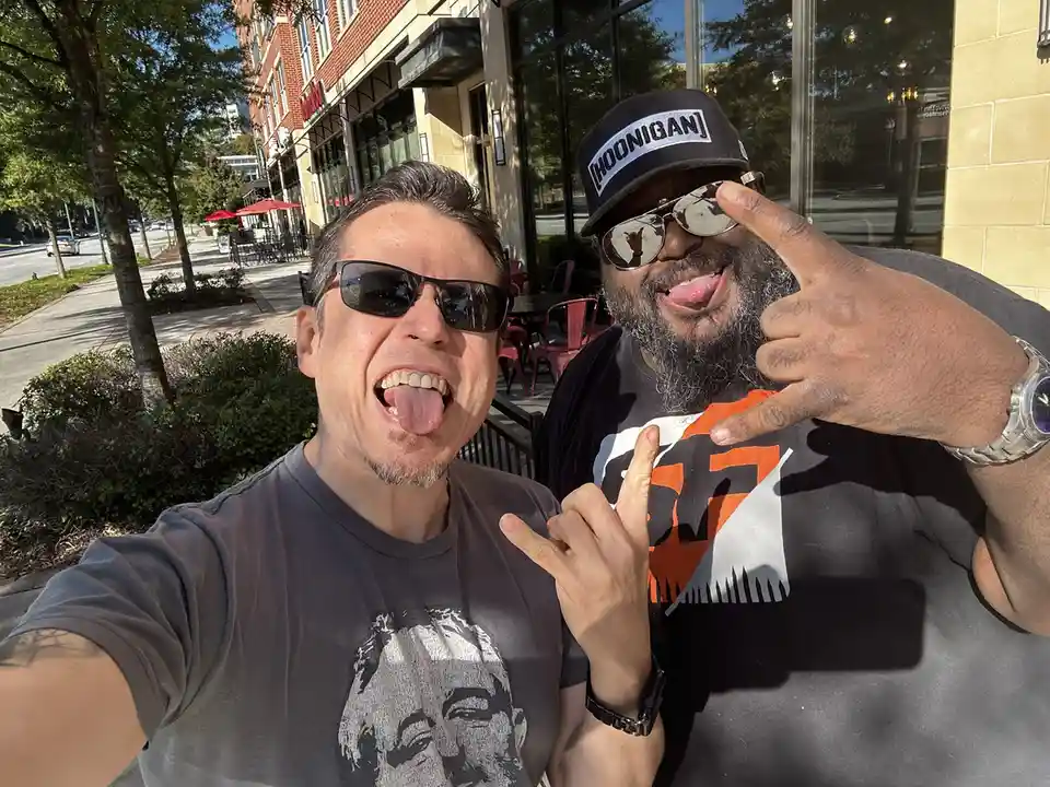 Two men pose for a selfie outdoors, both sticking out their tongues and making hand gestures. The man on the left, holding…