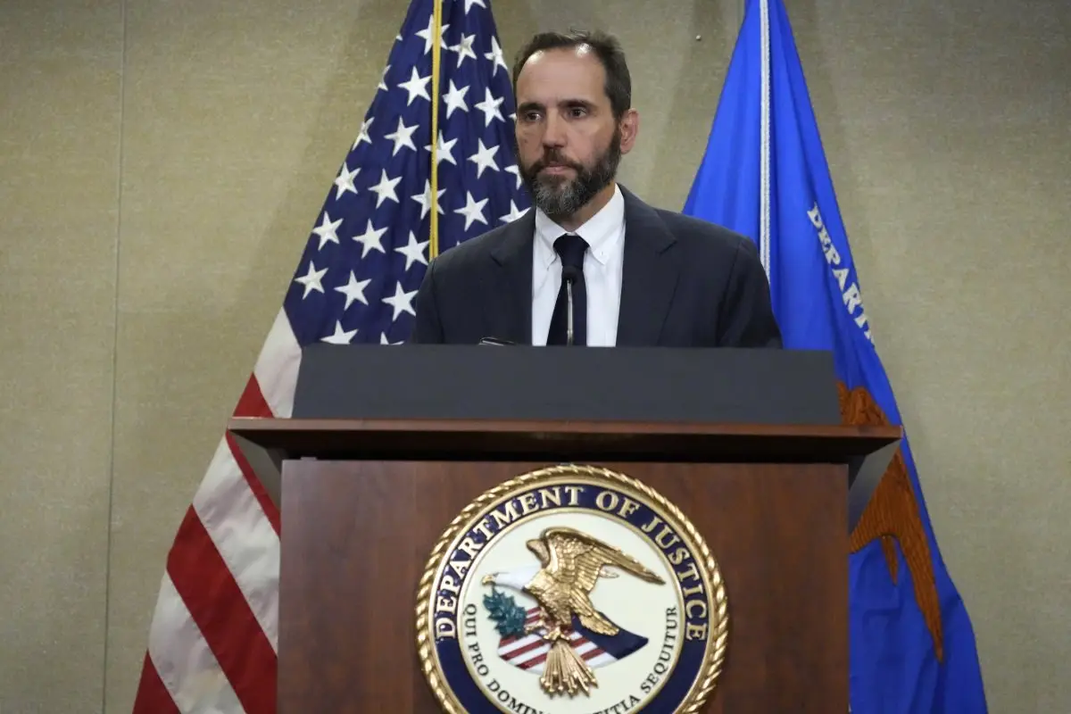 a man with a beard in a suit stands at a wooden podium bearing the Department of Justice seal, flanked by an american flag…