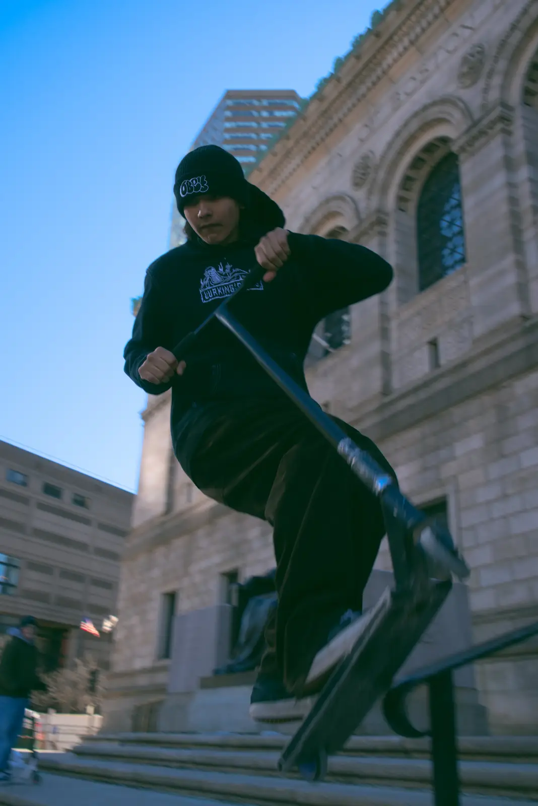 A person in all-black streetwear, including a beanie and hoodie, is mid-air performing a trick on a scooter while grinding a…