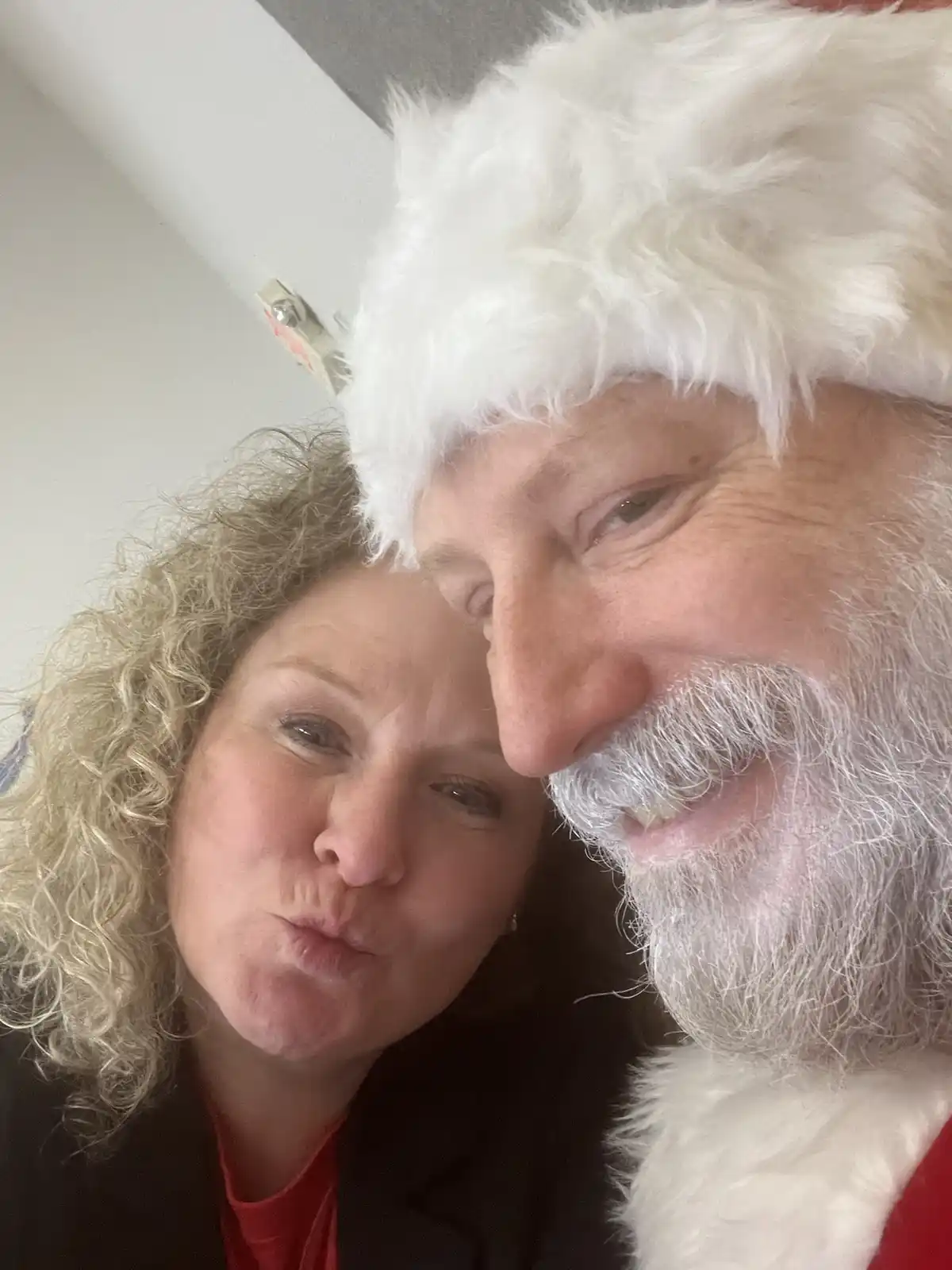 A close-up selfie shows a man dressed as Santa Claus with a white beard and hat, smiling warmly while his forehead touches a…