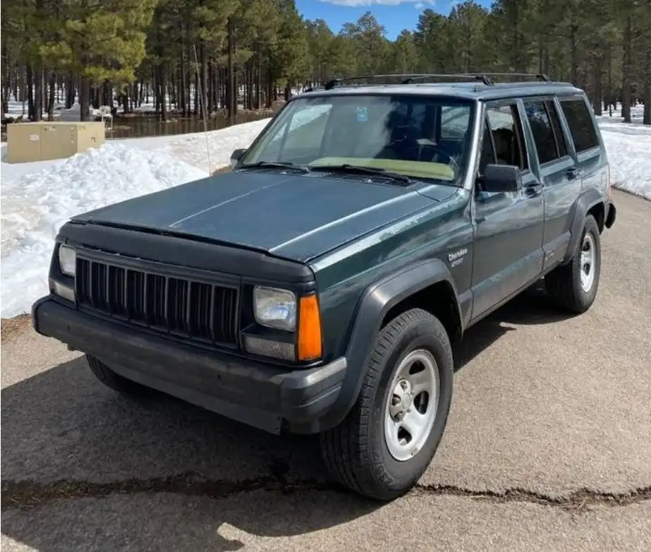 a dark green 1990s jeep cherokee station wagon is parked on a cracked asphalt road with snowbanks and pine trees in the…