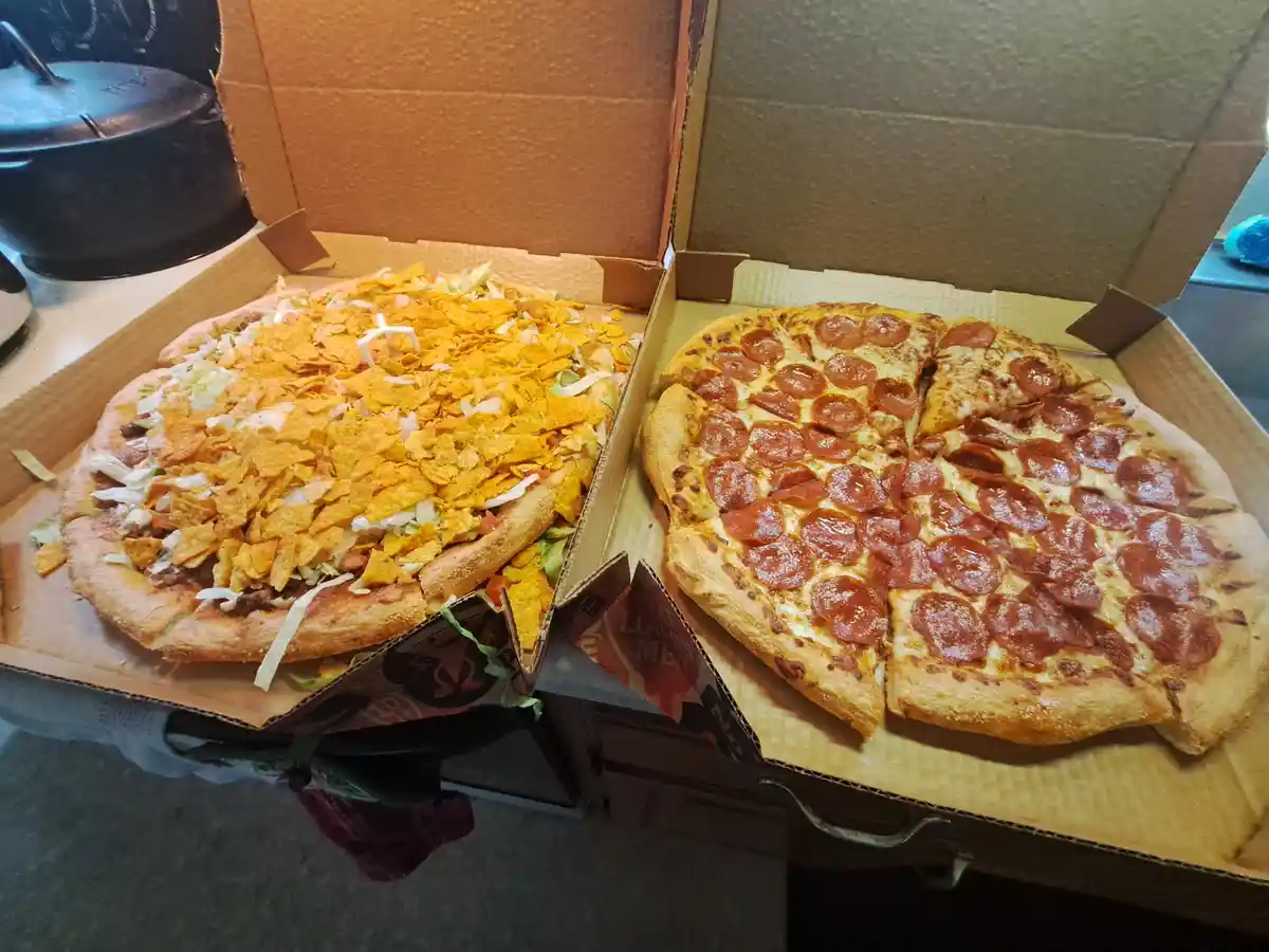 Two open pizza boxes sit side by side on a kitchen counter. The box on the left contains a pizza topped with orange tortilla…