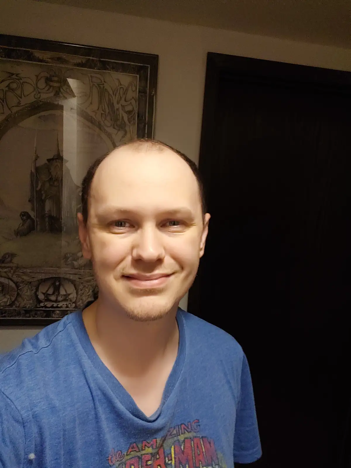 A man with a receding hairline and a slight smile wears a blue t-shirt with 'The Amazing Spider-Man' text. Behind him, a…