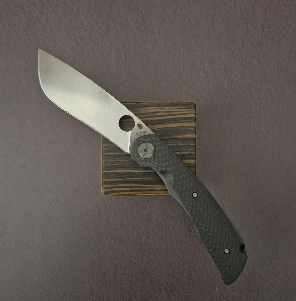A Spyderco brand folding knife with a brushed stainless steel blade and dark textured handle is displayed open on a small…