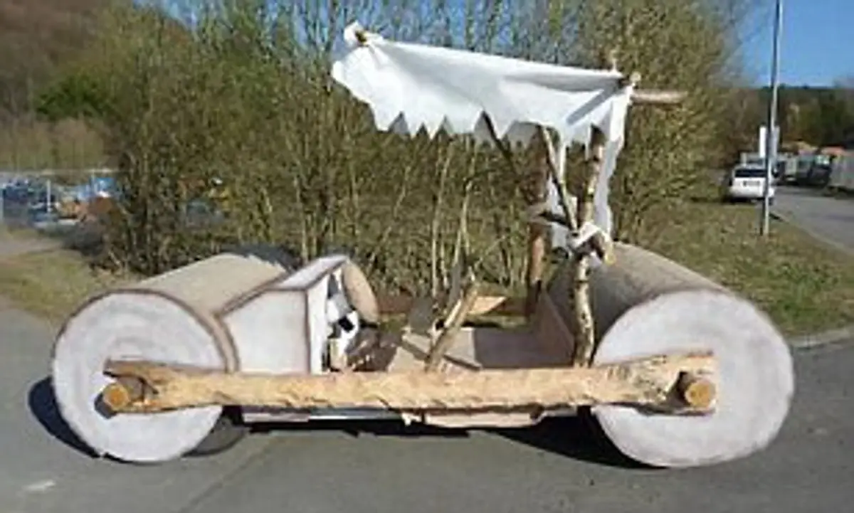 A crudely constructed vehicle sits on a paved road, featuring two massive, unfinished log wheels, a simple wooden frame, and…