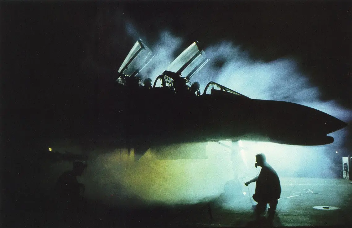 A dramatic nighttime photo shows a fighter jet with its canopy open, emitting smoke or steam, while a silhouetted person in…