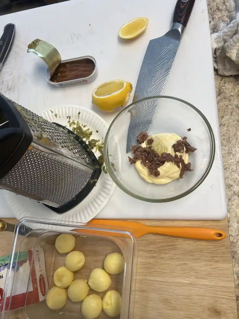 A kitchen counter displays ingredients for a recipe: a bowl of mayonnaise topped with chopped anchovies, a grater with green…