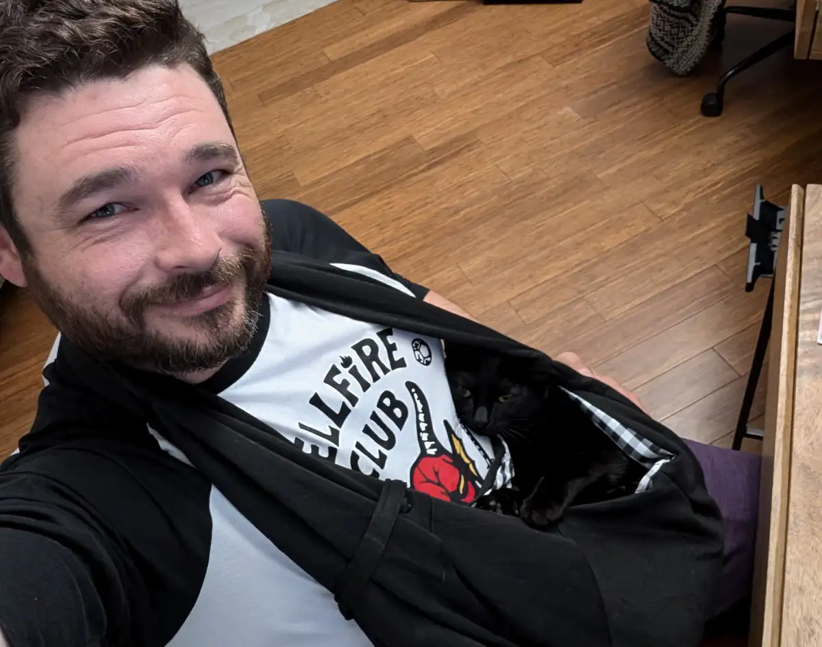 A man with a beard and blue eyes smiles at the camera while holding open his black jacket to reveal a black cat nestled…