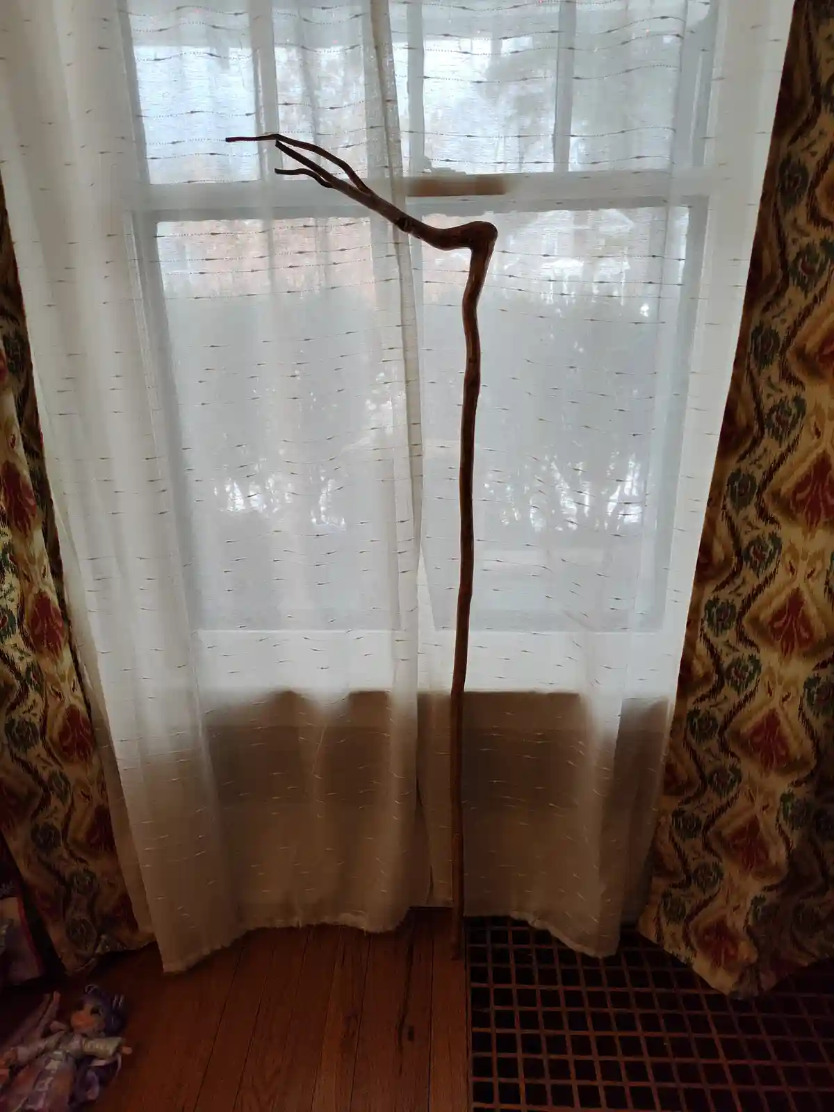 A long, gnarled wooden stick stands upright in front of a sheer white curtain, its top end split into three forked branches…