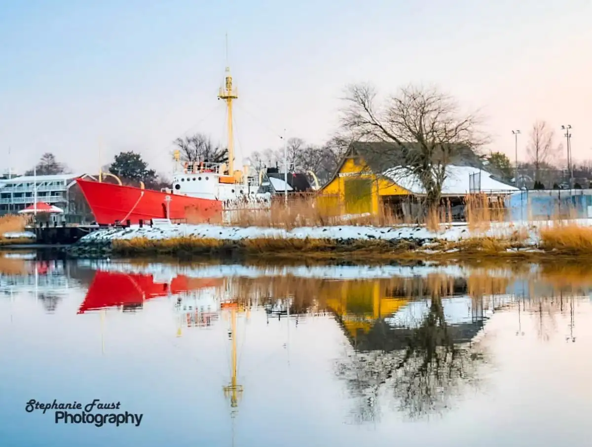 A serene winter scene shows a red boat and yellow building reflected in calm water, with snow on the ground and bare trees…