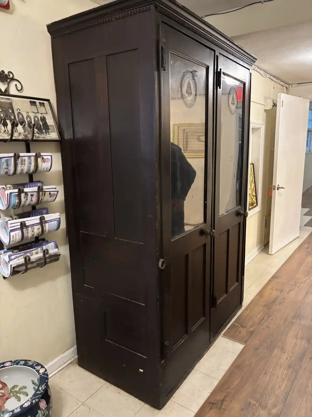 A tall, dark wooden cabinet with glass-paneled doors stands in a hallway, featuring a circular 'public telephone' logo on…