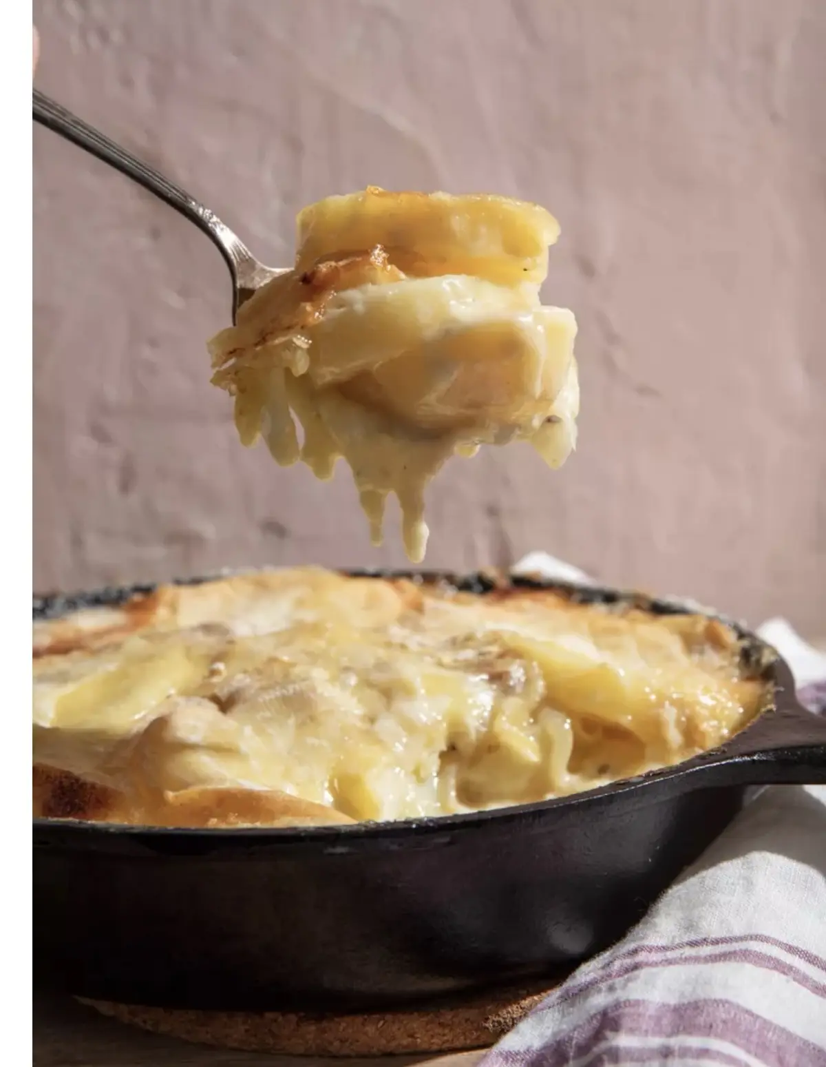 A close-up photo shows a fork lifting a generous, gooey scoop of baked macaroni and cheese from a black cast-iron skillet…