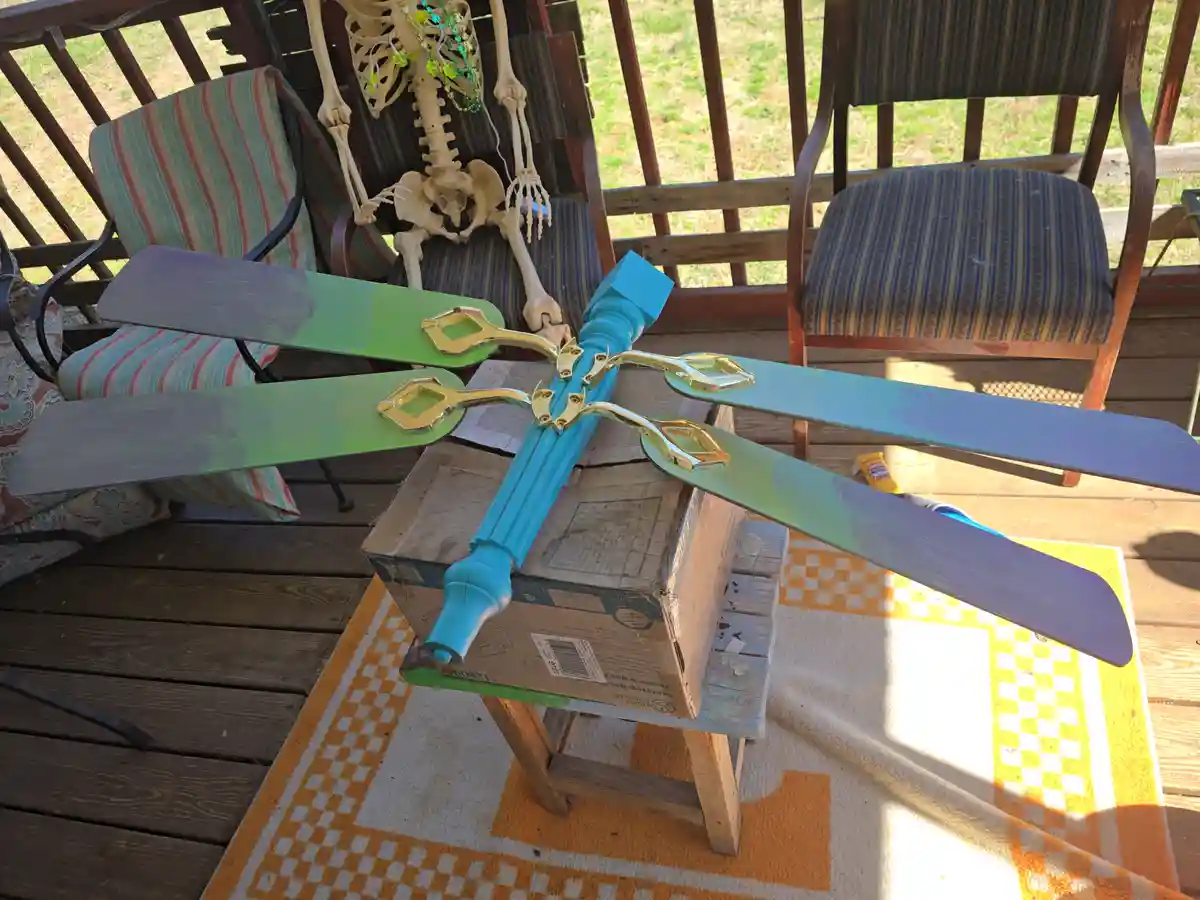 A ceiling fan with blades painted in a gradient of green and blue, resting on a cardboard box on a porch. In the background…
