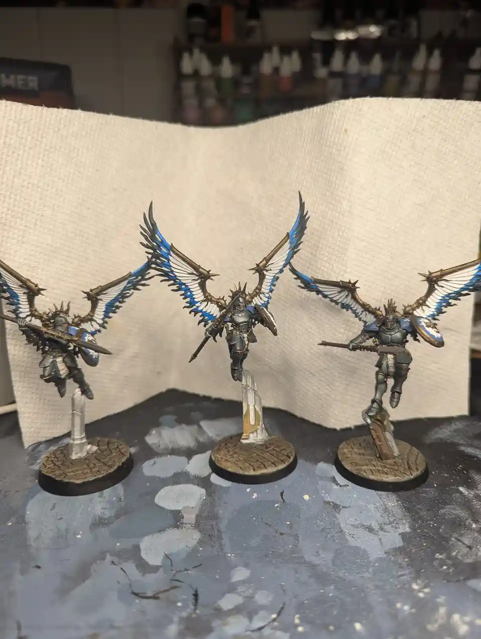 Three intricately painted miniature fantasy warriors with large, spread wings in blue, white, and gold stand on bases…