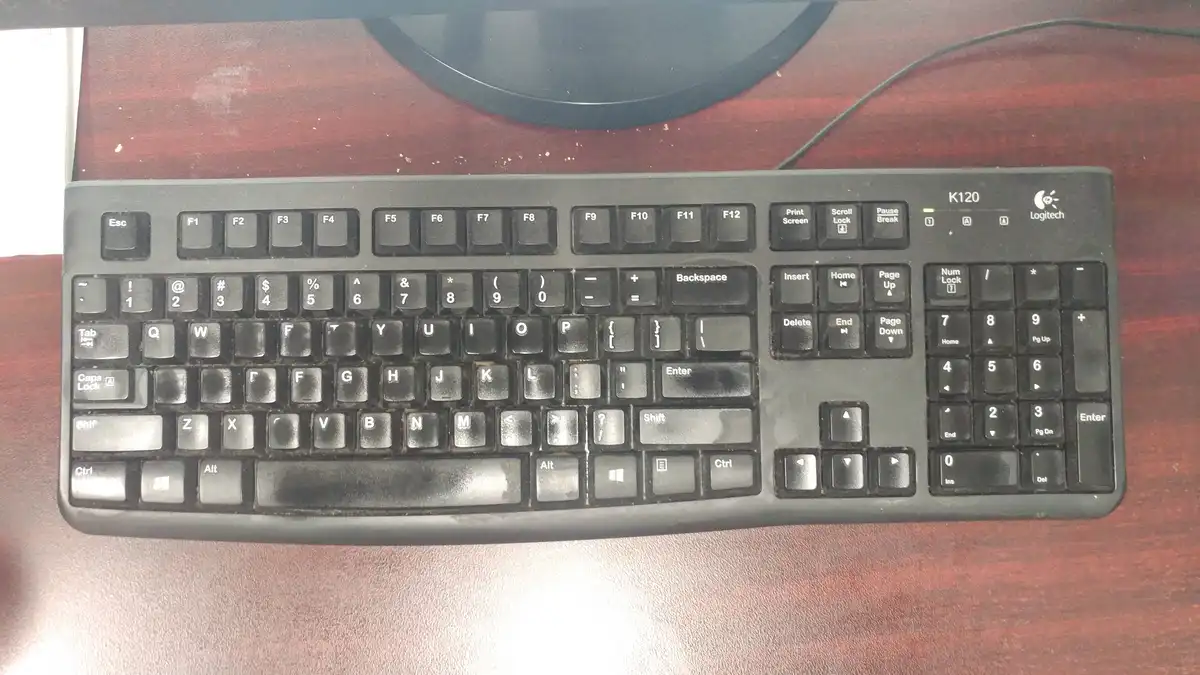 a black logitech k120 keyboard with visibly worn and dirty keys sits on a wooden desk, with a monitor base visible above it…