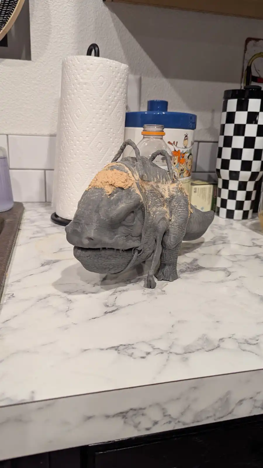 A grey plastic dinosaur figurine sits on a white marble kitchen counter, covered in a layer of light brown dust…