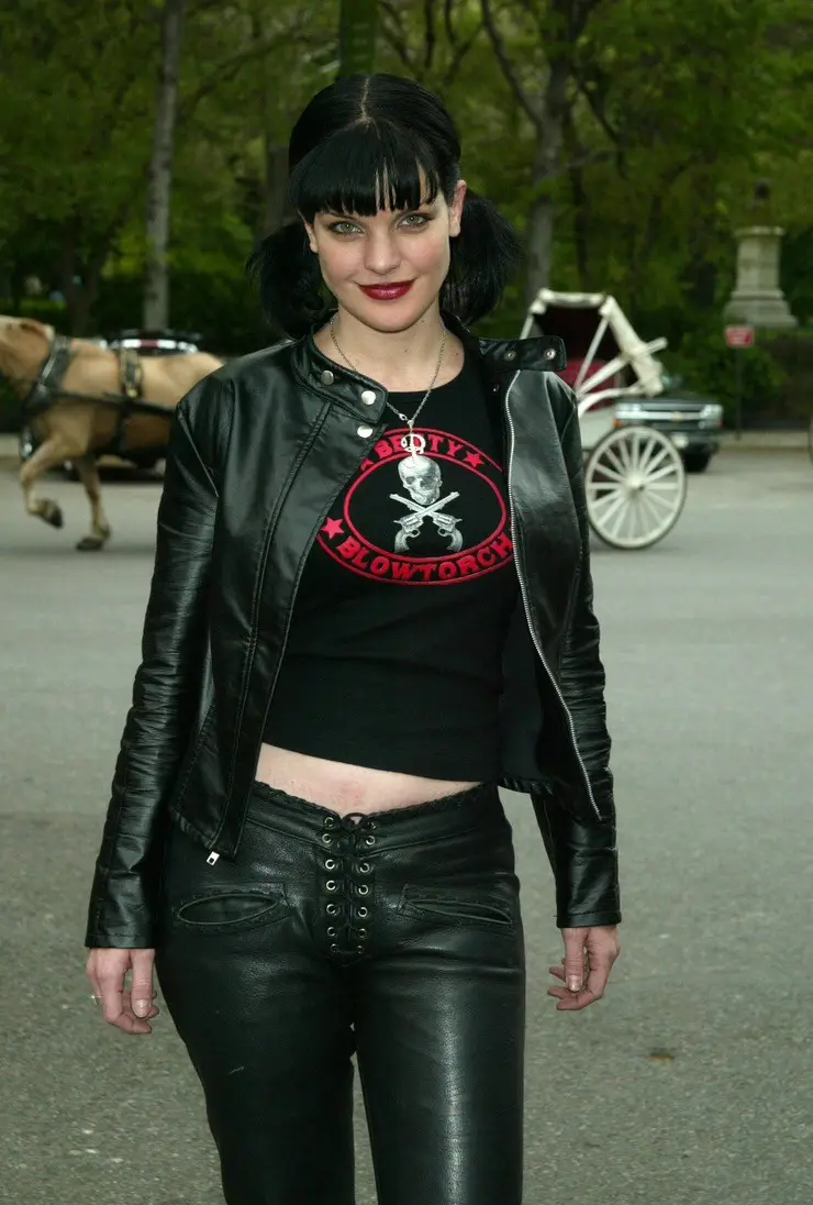 A woman with black hair and bangs, wearing a black leather jacket over a cropped black t-shirt with a red circular logo…