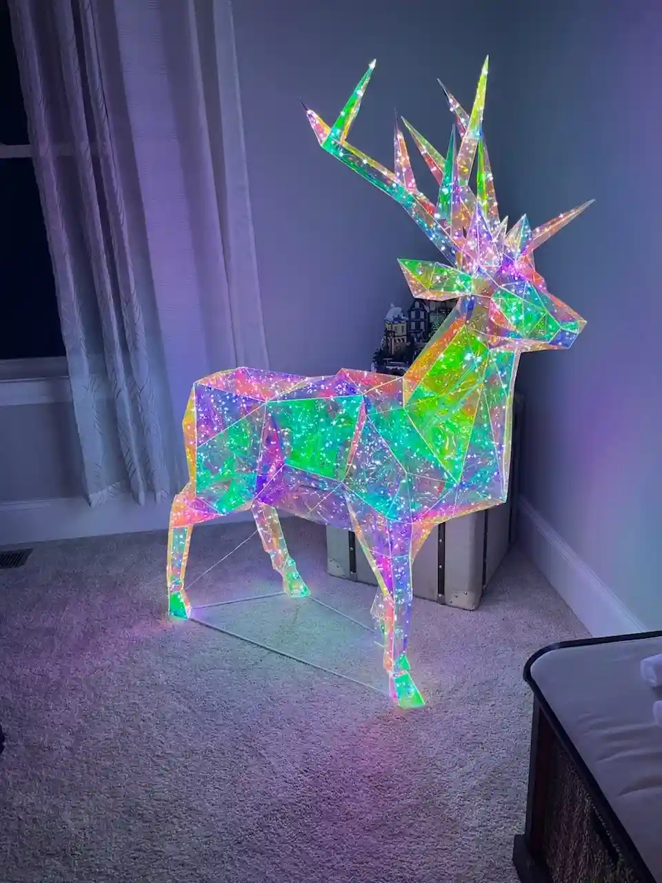 a large, geometric, illuminated deer sculpture stands in a room, its body and antlers made of glowing, multicolored panels…